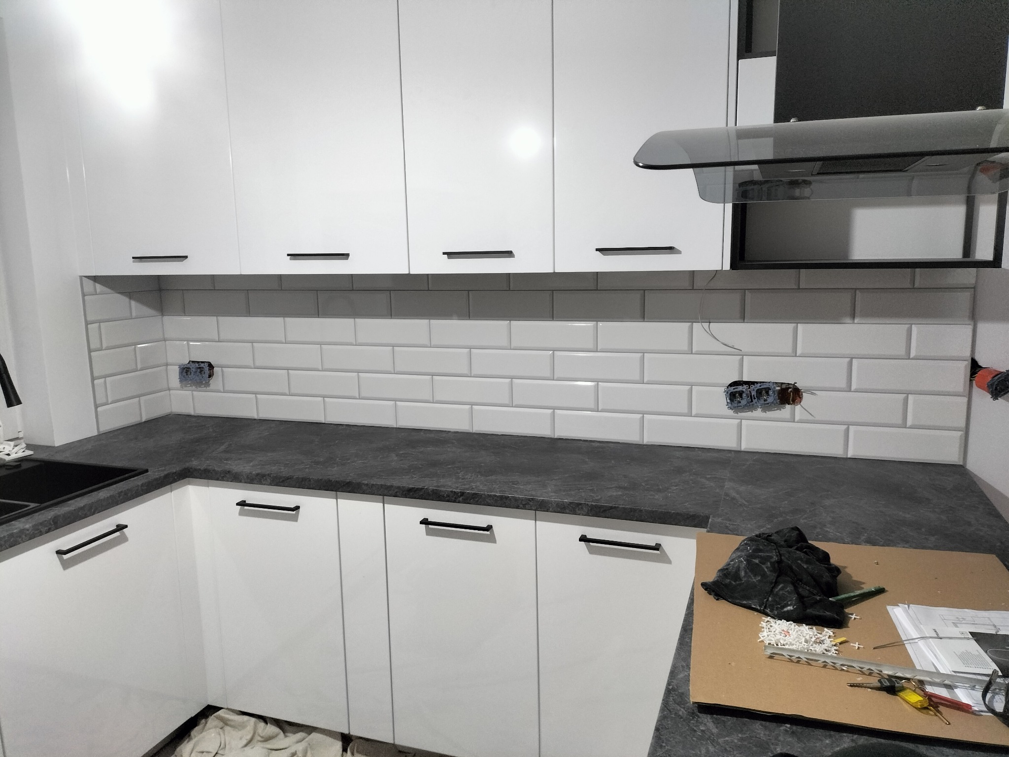 Biała kuchnia z czarnymi uchwytami i blatem, wykończona białymi płytkami typu metro. Widoczne gniazdka elektryczne i narzędzia na blacie.