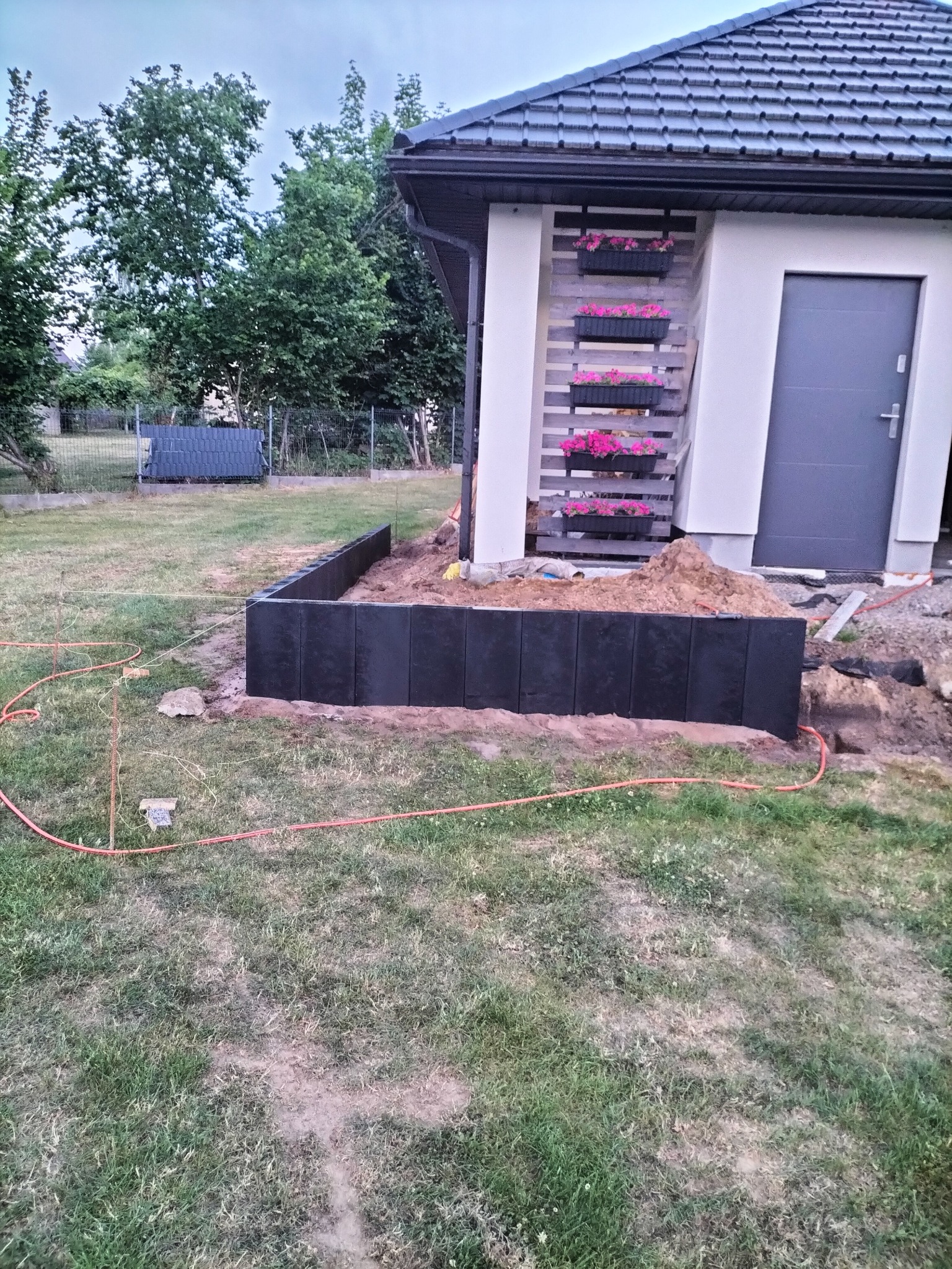 Częściowo ukończona rabata z ciemnych, betonowych elementów obok budynku z kwiatami w donicach. Widoczne narzędzia i ziemia. Prace w toku.