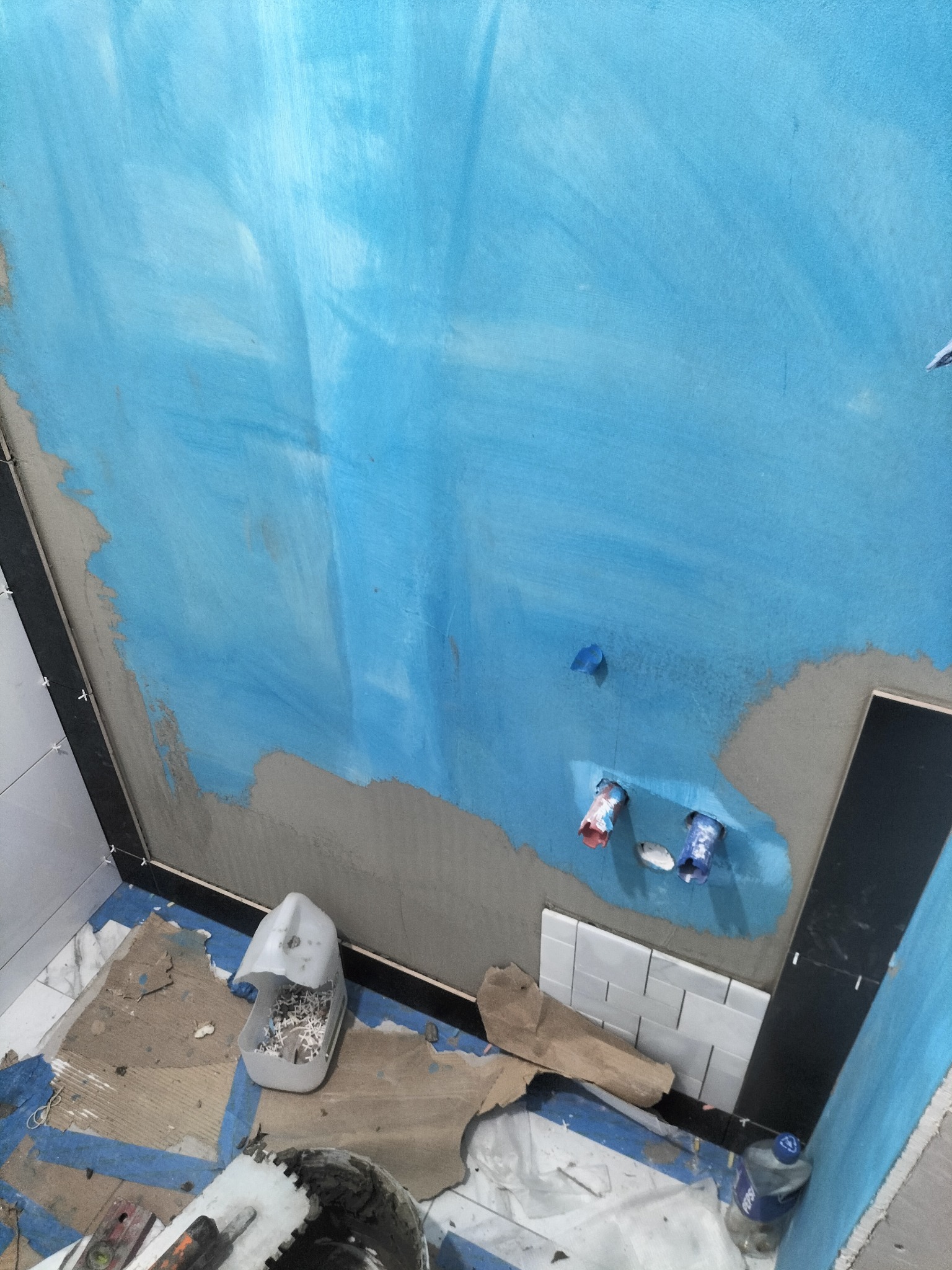 Niebieska hydroizolacja na ścianie łazienki w trakcie remontu, widoczne rury, fragment ułożonych białych płytek i narzędzia glazurnicze na podłodze. Ujęcie z dołu.