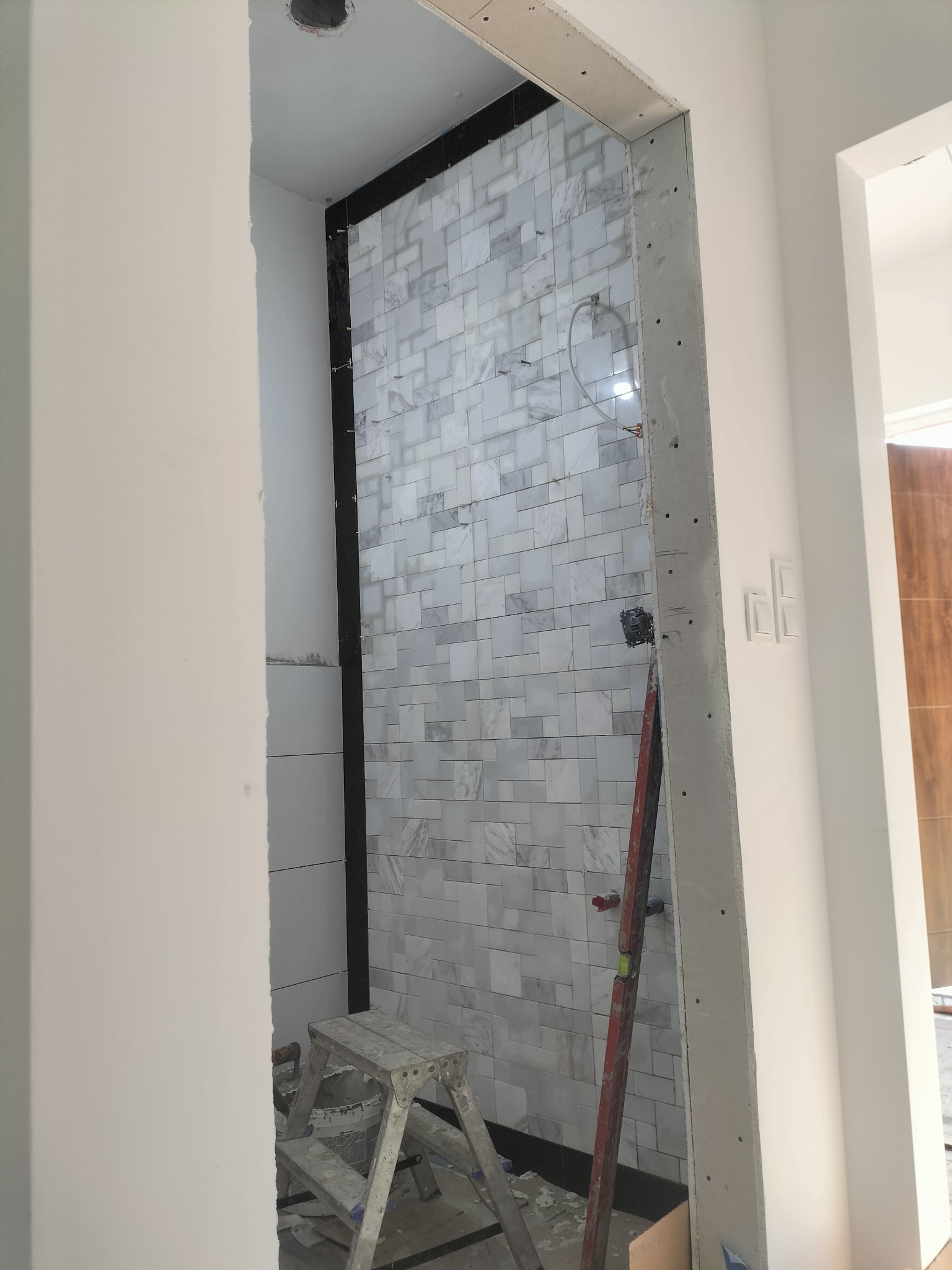 Wnętrze łazienki w trakcie wykańczania: białe marmurowe płytki na ścianie, drabina malarska, poziomica oparta o ścianę, widoczne przewody elektryczne.