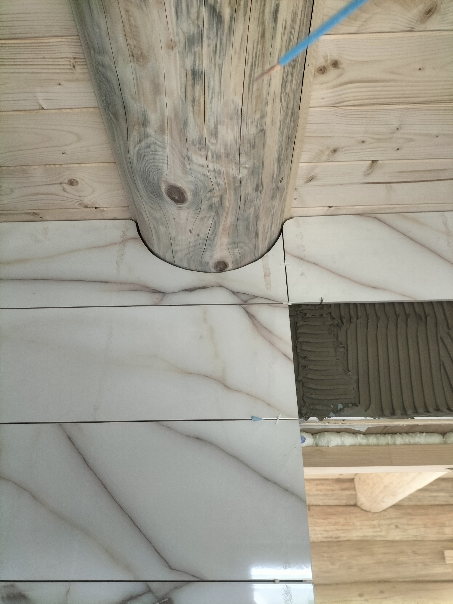 Układanie marmurowych płytek ściennych wokół drewnianego słupa w rustykalnym wnętrzu. Widoczna zaprawa i niebieski przewód elektryczny.