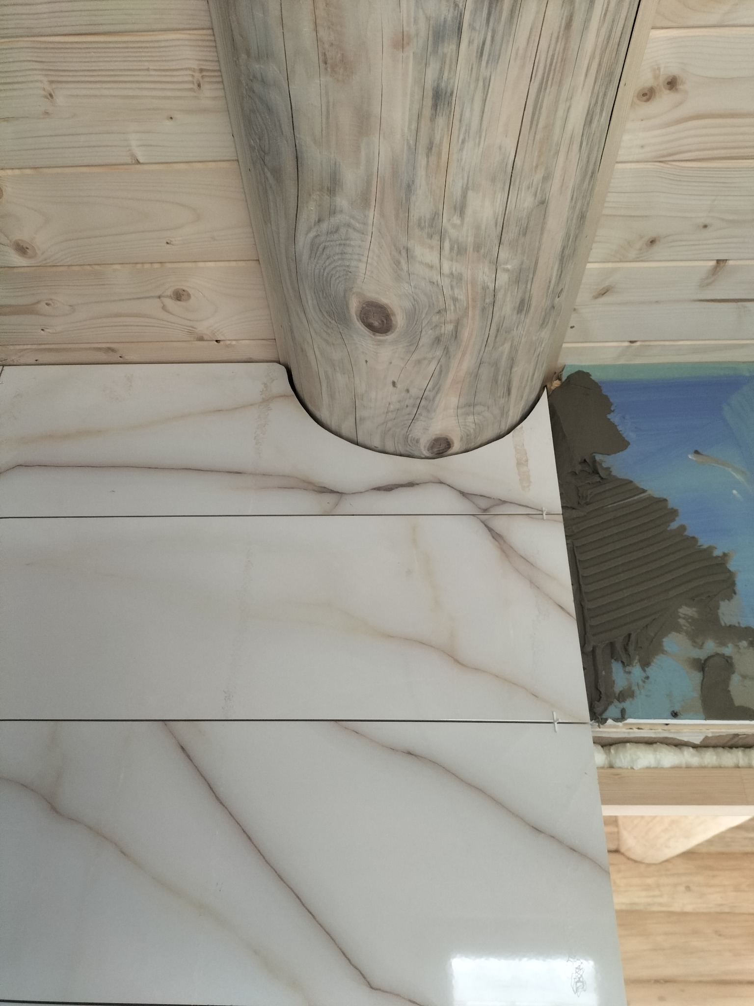 Układanie płytek imitujących marmur wokół drewnianego słupa w domu z bali. Widoczna zaprawa i krzyżyki dystansowe. Detal wykończenia wnętrza.