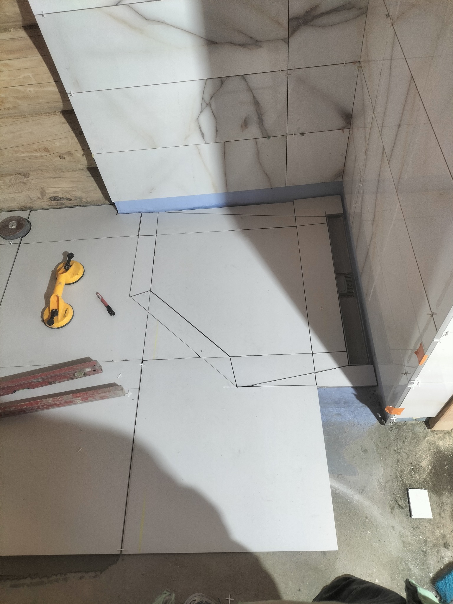 Układanie białych, marmurkowych płytek w łazience, widoczny odpływ liniowy, przyssawka do płytek i poziomica. Etap prac wykończeniowych, widoczne fugi i krzyżyki dystansowe.