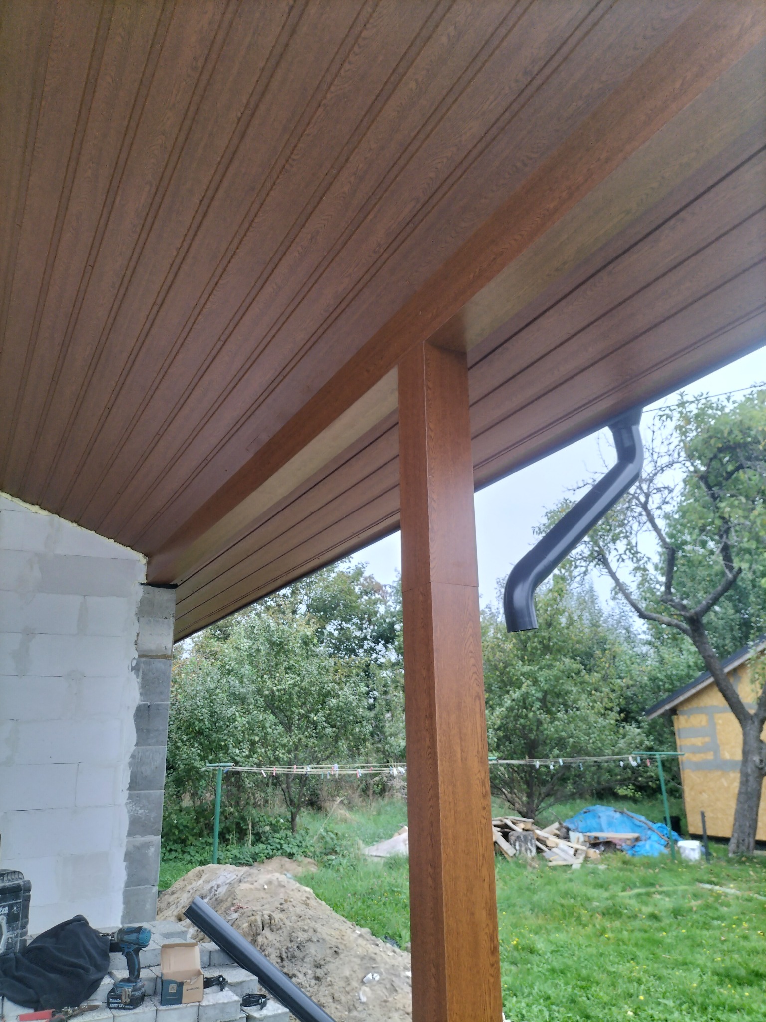 Wykończony podbitka dachowa z brązowego panelu PCV, widoczna kolumna wspierająca i fragment orynnowania. W tle budynek w trakcie budowy i zieleń.