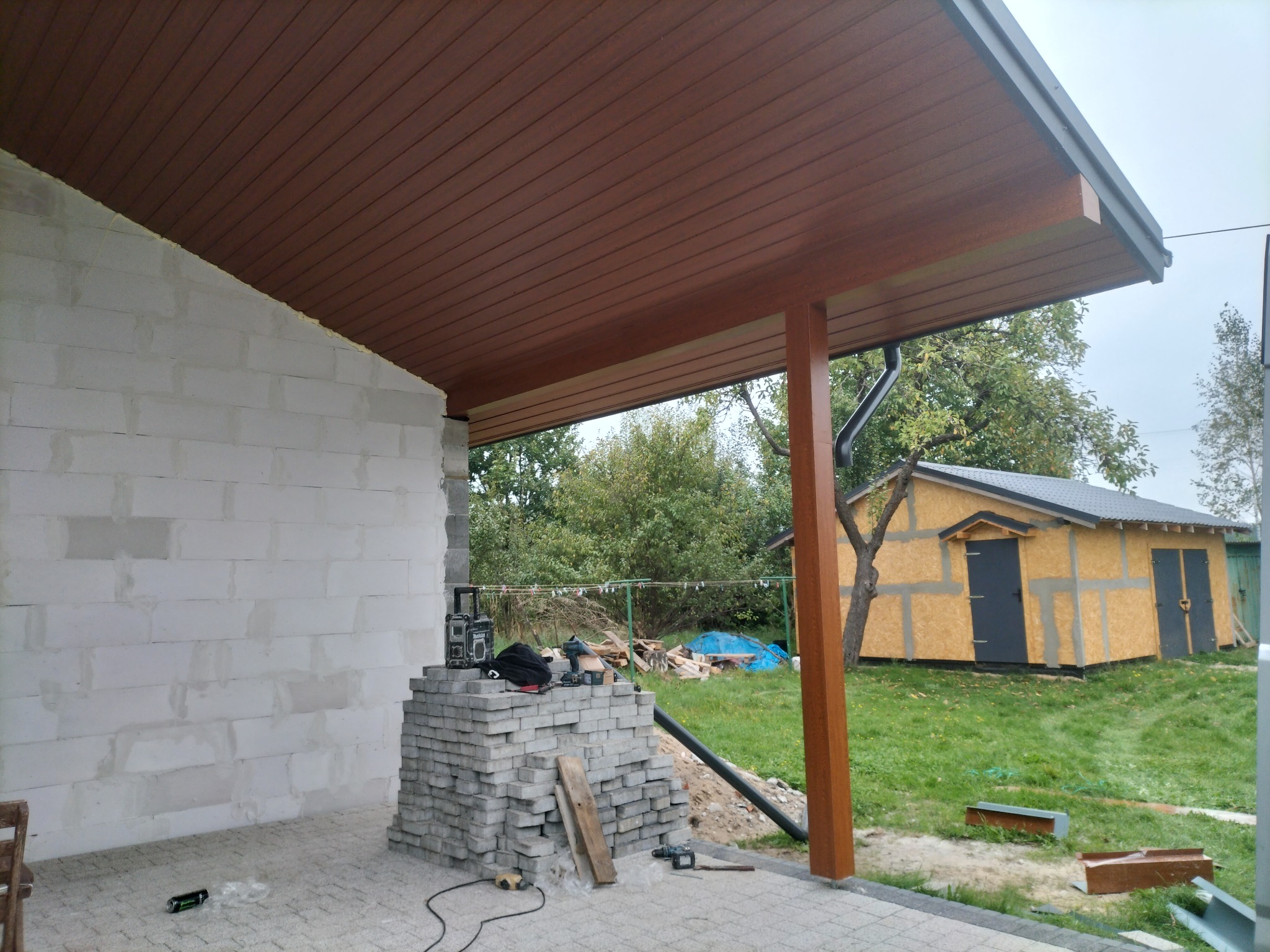 Zadaszenie tarasu w trakcie budowy: brązowe panele podsufitki, białe ściany z bloczków, narzędzia na palecie. W tle drewniany budynek gospodarczy i zieleń.
