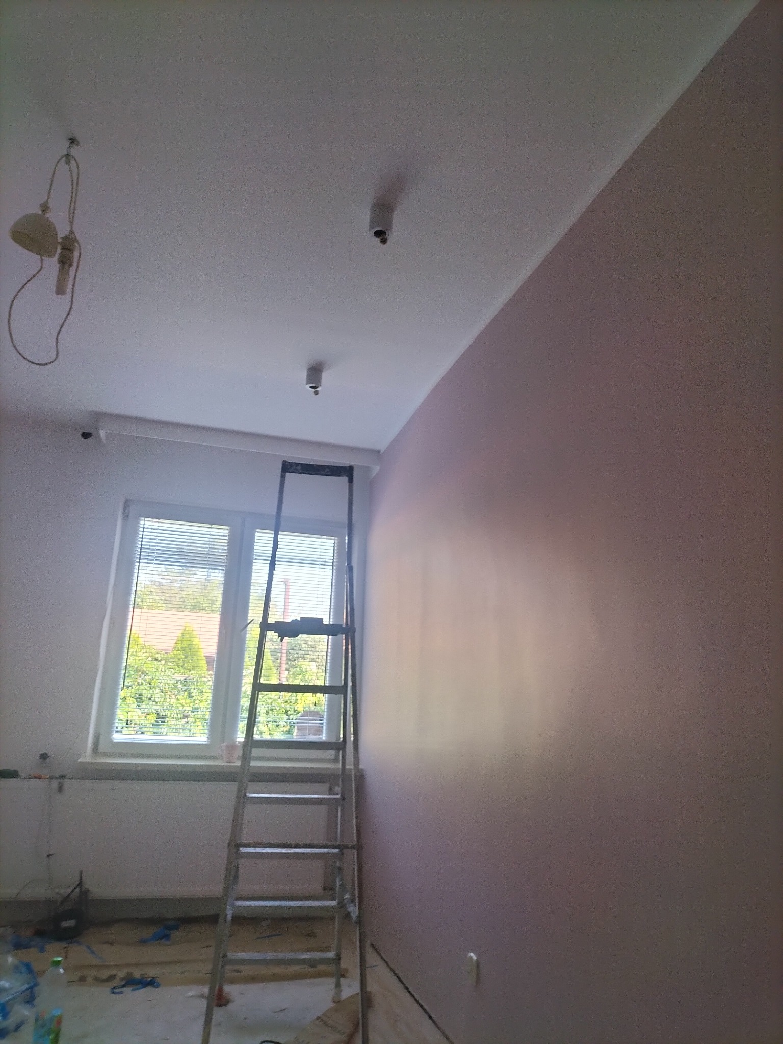 Pokój w trakcie malowania na różowo, drabina oparta o ścianę, widoczne niedoskonałości i resztki po remoncie, okno z żaluzjami w tle.