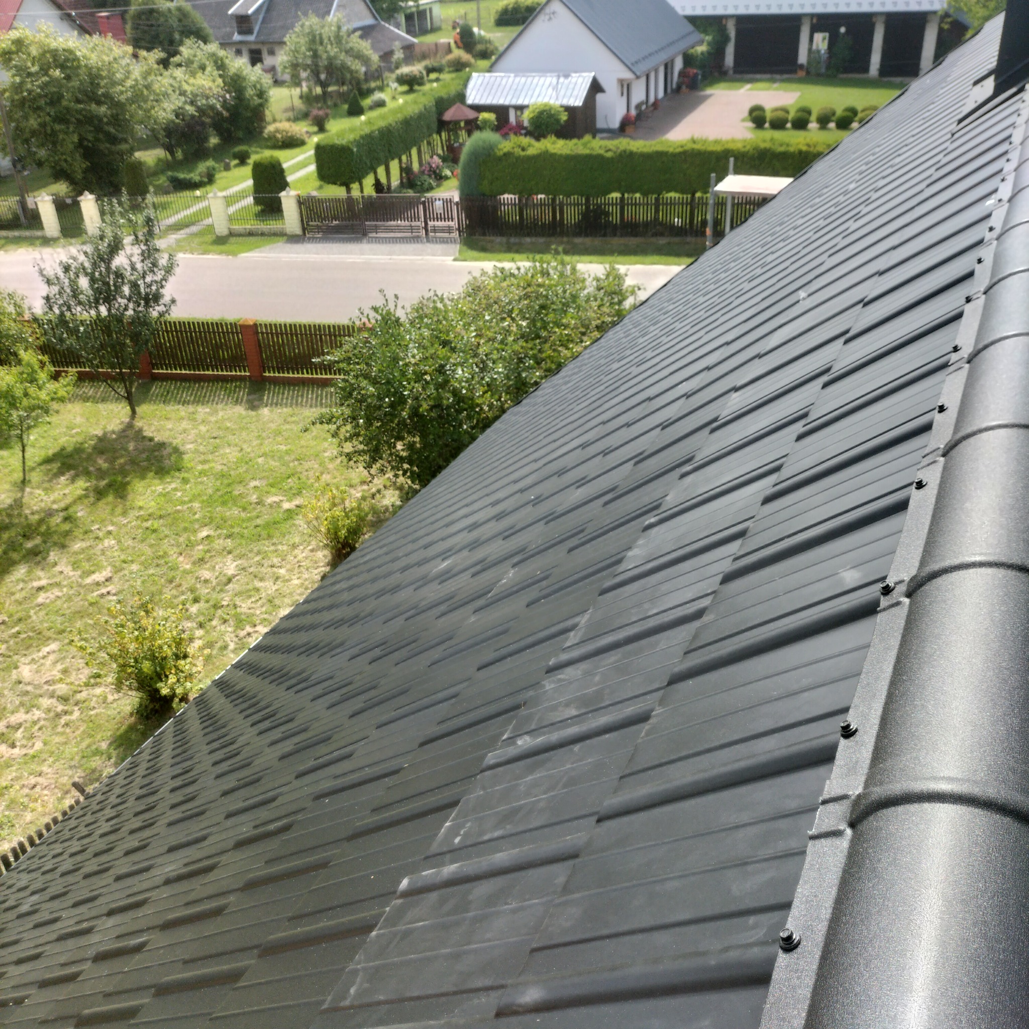 Szczegółowe ujęcie grafitowego dachu z blachy trapezowej, widok z góry na domy i zieleń w tle, mocowania widoczne.