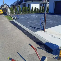 Nowoczesny podjazd z ciemnoszarej kostki brukowej ułożonej w kwadraty, obrzeża z jasnych płyt, w tle dom z ciemną bramą garażową i żółty samochód dostawczy, widoczne narzędzia i maszyna...