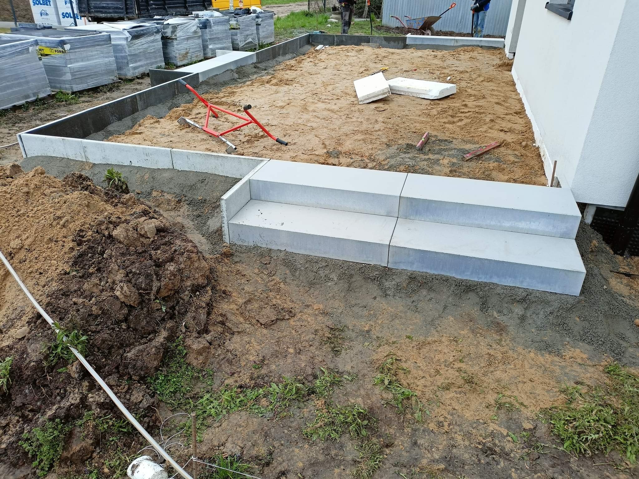 Przygotowanie podłoża pod kostkę brukową z widocznymi obrzeżami, schodami z kostki i stosem kostki Solbet w tle.