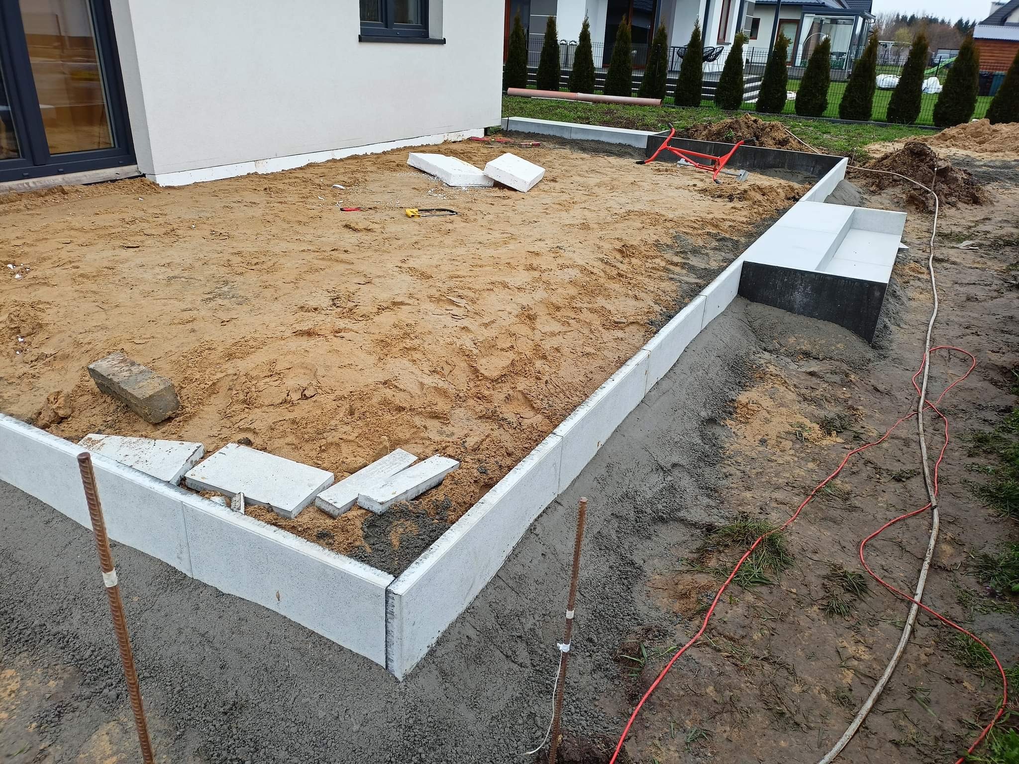 Przygotowanie terenu pod taras: ułożone obrzeża z jasnoszarej kostki betonowej, podsypka piaskowa, wystające pręty zbrojeniowe i rozłożone kable elektryczne na obrzeżach działki.