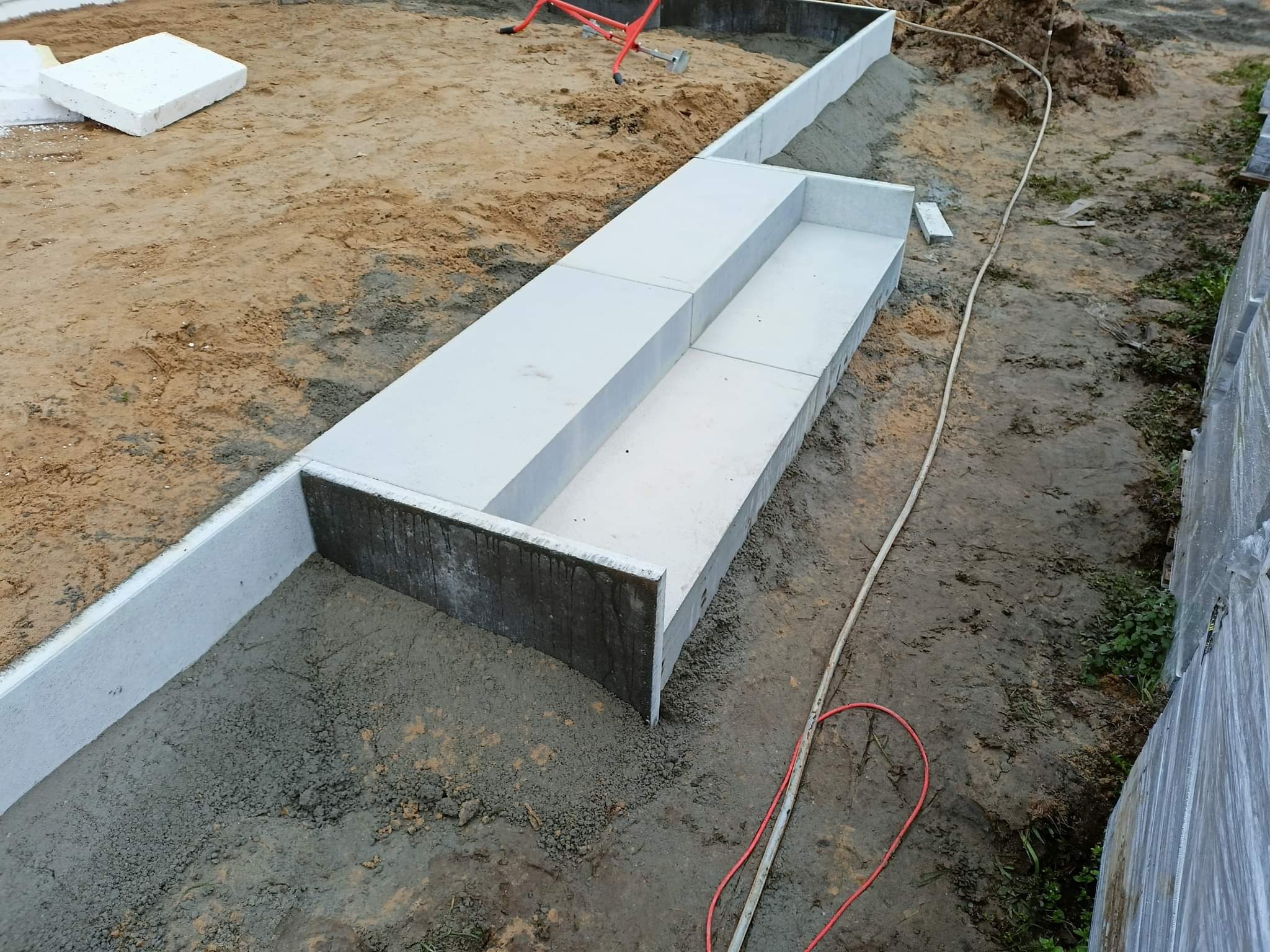 Betonowe schody zewnętrzne w trakcie budowy, widoczne szare elementy konstrukcyjne i ziemiste podłoże, czerwony przewód na ziemi, etap prac wykończeniowych.