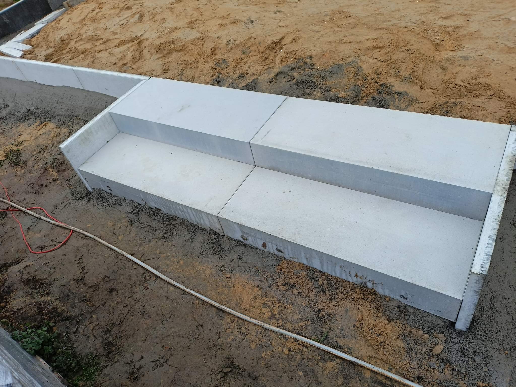 Dwu-stopniowe schody z betonowych płyt, w trakcie budowy, widoczne przewody elektryczne i przygotowane podłoże z piasku i żwiru.