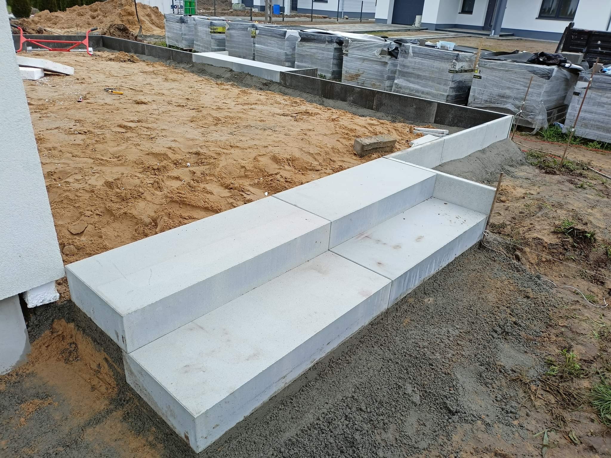 Przygotowanie podłoża pod schody zewnętrzne z betonowych bloków, widoczny piasek, podsypka cementowo-piaskowa i palety z kostką brukową w tle.