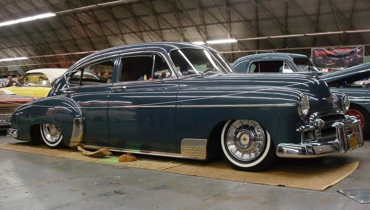 Niebieski, obniżony klasyczny samochód na wystawie, z chromowanymi detalami i białymi oponami, zaparkowany na brązowej macie w hali.