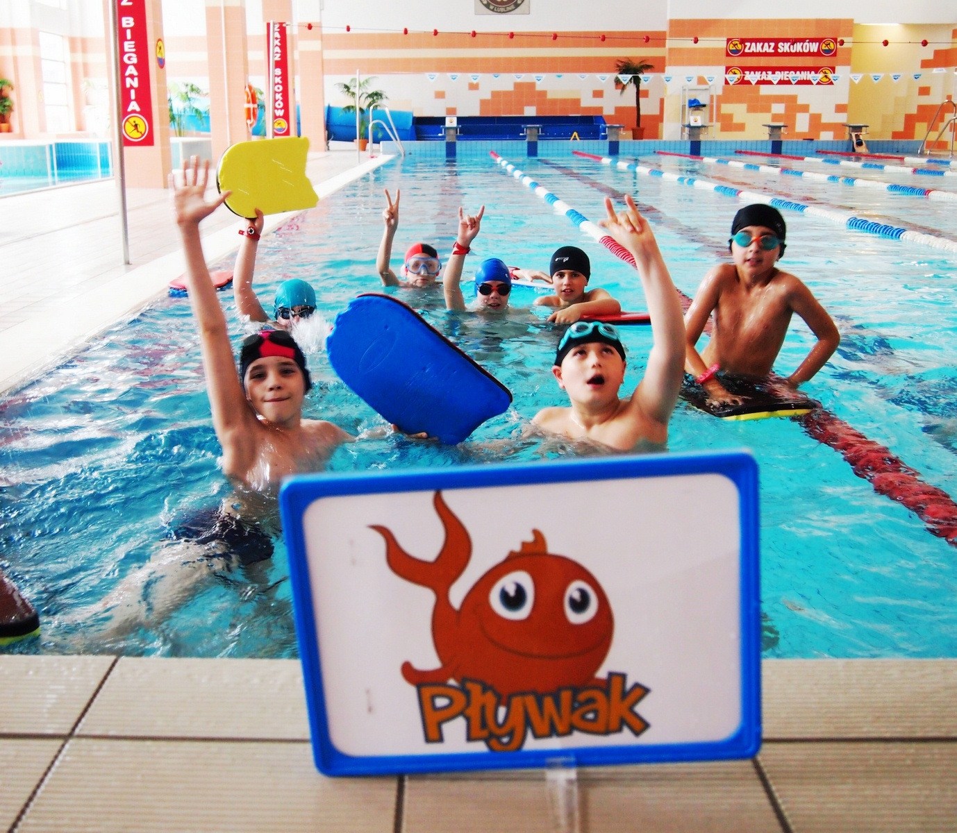 Grupa dzieci w basenie, część z deskami do pływania, machają rękami, w pierwszym planie tabliczka z rysunkiem pomarańczowej rybki i napisem 'Pływak'.