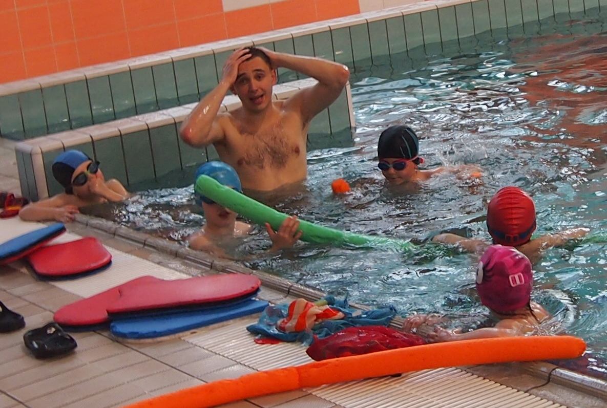 Instruktor z podopiecznymi w basenie, dzieci w czepkach i okularach pływackich, jedno trzyma zielonego makarona, na brzegu leżą deski i ręczniki.