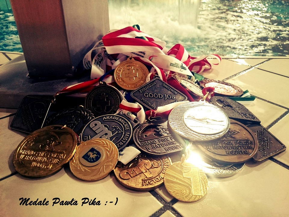 Kolekcja medali pływackich z różnymi inskrypcjami i wstążkami, ułożona na kafelkach przy fontannie, z widocznym napisem 'Medale Pawła Pika'.