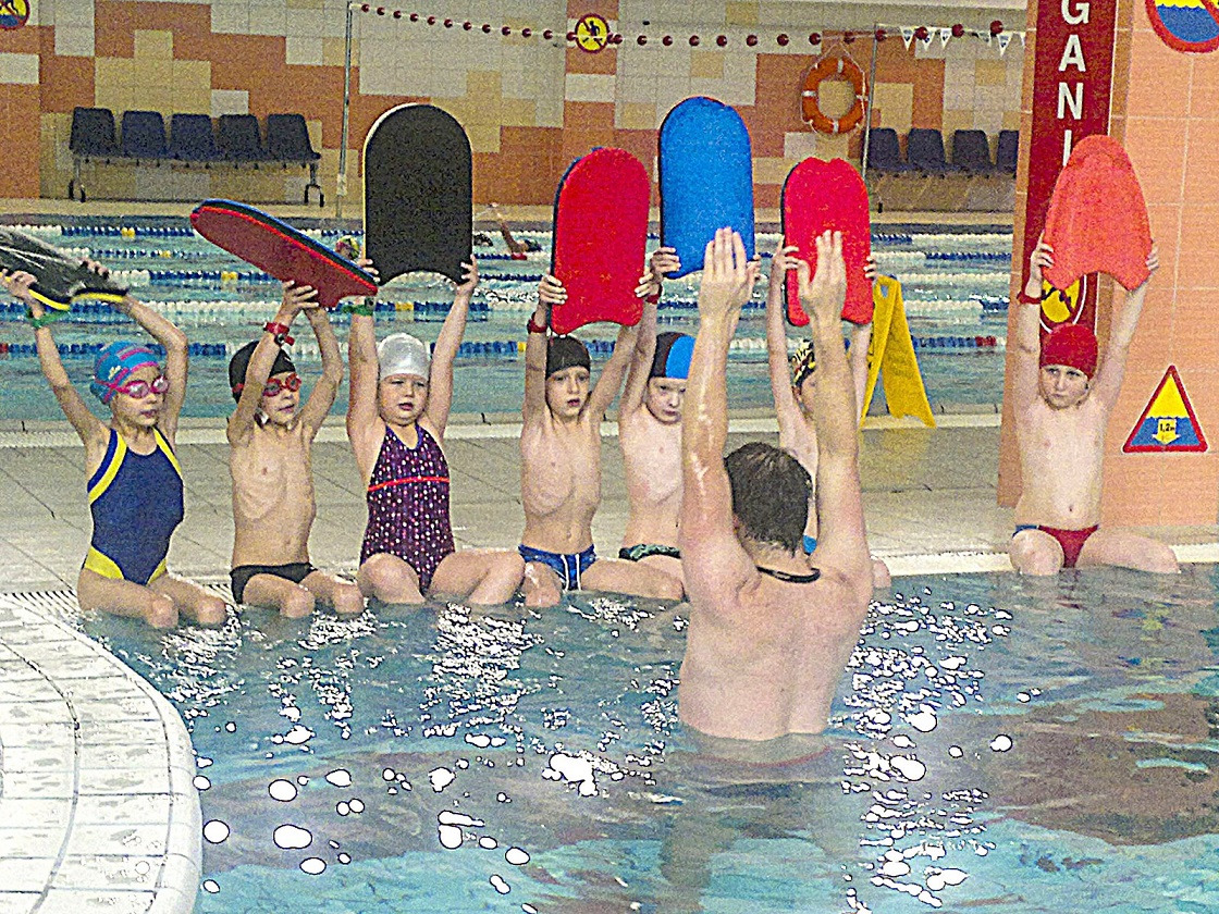 Grupa dzieci siedzi na brzegu basenu podczas lekcji pływania, trzymając deski do pływania nad głowami, pod nadzorem instruktora.