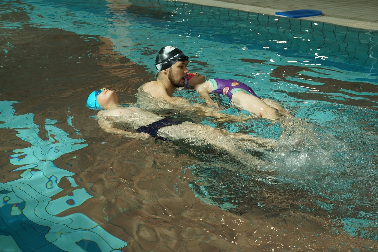 Instruktor w czepku z dwójką dzieci unoszących się na wodzie w basenie, podczas lekcji pływania, odbicia światła na dnie basenu.