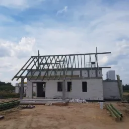 Budowa domu jednorodzinnego w stanie surowym otwartym, widoczne ściany z bloczków, drewniana konstrukcja dachu w trakcie montażu, materiały budowlane na placu budowy.