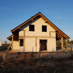 Surowy stan domu jednorodzinnego z pustaków, z widoczną konstrukcją dachu, bez okien i drzwi, z żółtą rurą wentylacyjną leżącą na ziemi, w tle kościół z wieżą zwieńczoną krzyżem.