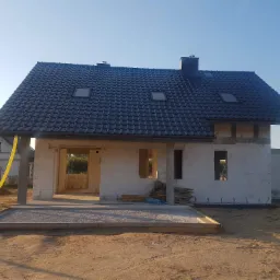 Dom w trakcie budowy z czarną dachówką, widoczne okna dachowe i komin, ściany z bloczków, żółte rury wentylacyjne na zewnątrz budynku.