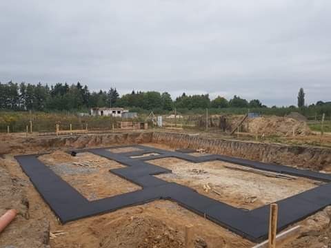 Przygotowanie fundamentów pod budowę domu jednorodzinnego: wykopane rowy fundamentowe z ułożoną izolacją poziomą, widoczne drewniane pale wyznaczające obrys budynku, w tle zarośnięta działka...