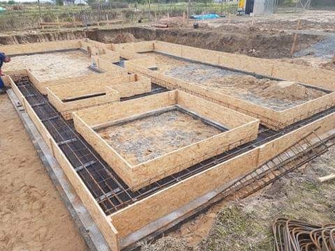 Przygotowanie szalunków i zbrojenia pod fundament domu jednorodzinnego na piaszczystym podłożu, widoczne drewniane formy i stalowe pręty zbrojeniowe.