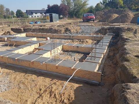 Fundamenty domu w trakcie budowy: szalunki z płyty OSB wypełnione świeżym betonem, zbrojenie pionowe, wykop w piaszczystej ziemi.