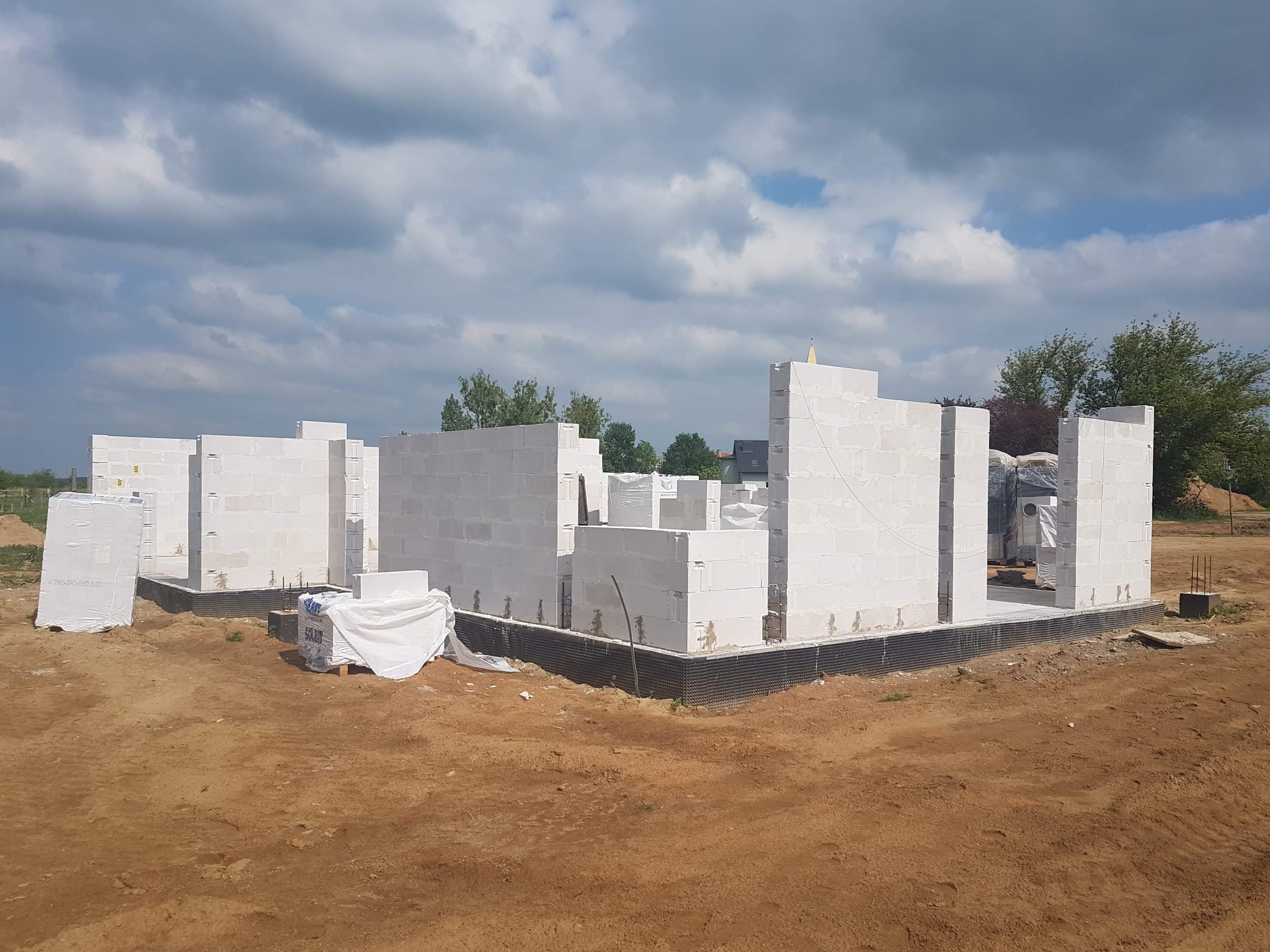 Wznoszenie ścian parterowego domu z białych bloczków na fundamencie z izolacją przeciwwilgociową, widoczne paczki materiałów budowlanych na ziemi i pochmurne niebo.