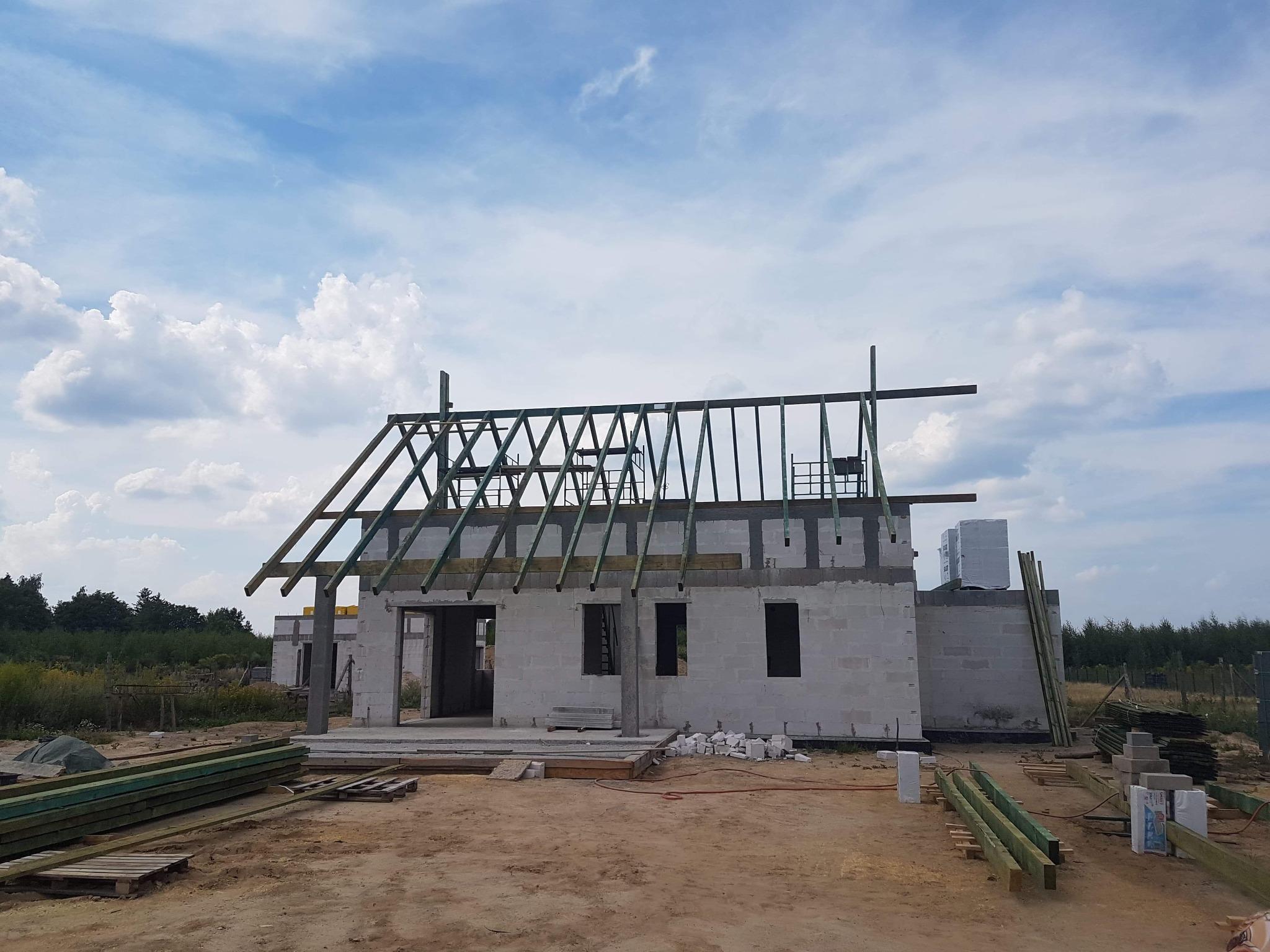 Budowa domu jednorodzinnego w stanie surowym otwartym, widoczne ściany z bloczków, drewniana konstrukcja dachu w trakcie montażu, materiały budowlane na placu budowy.