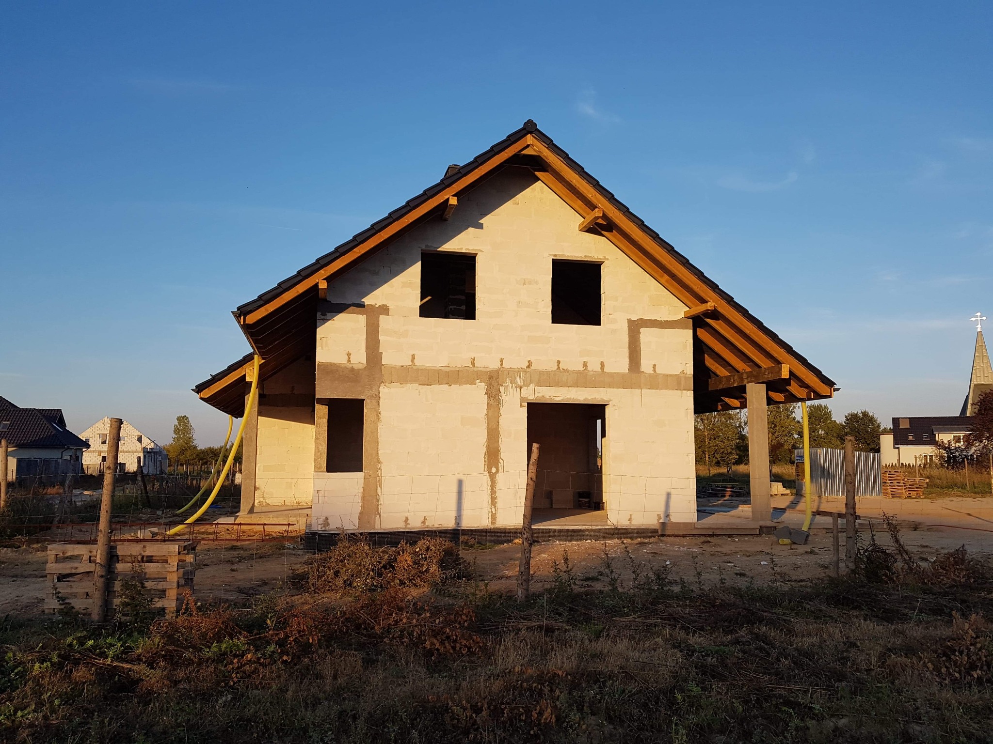 Surowy stan domu jednorodzinnego z pustaków, z widoczną konstrukcją dachu, bez okien i drzwi, z żółtą rurą wentylacyjną leżącą na ziemi, w tle kościół z wieżą zwieńczoną krzyżem.
