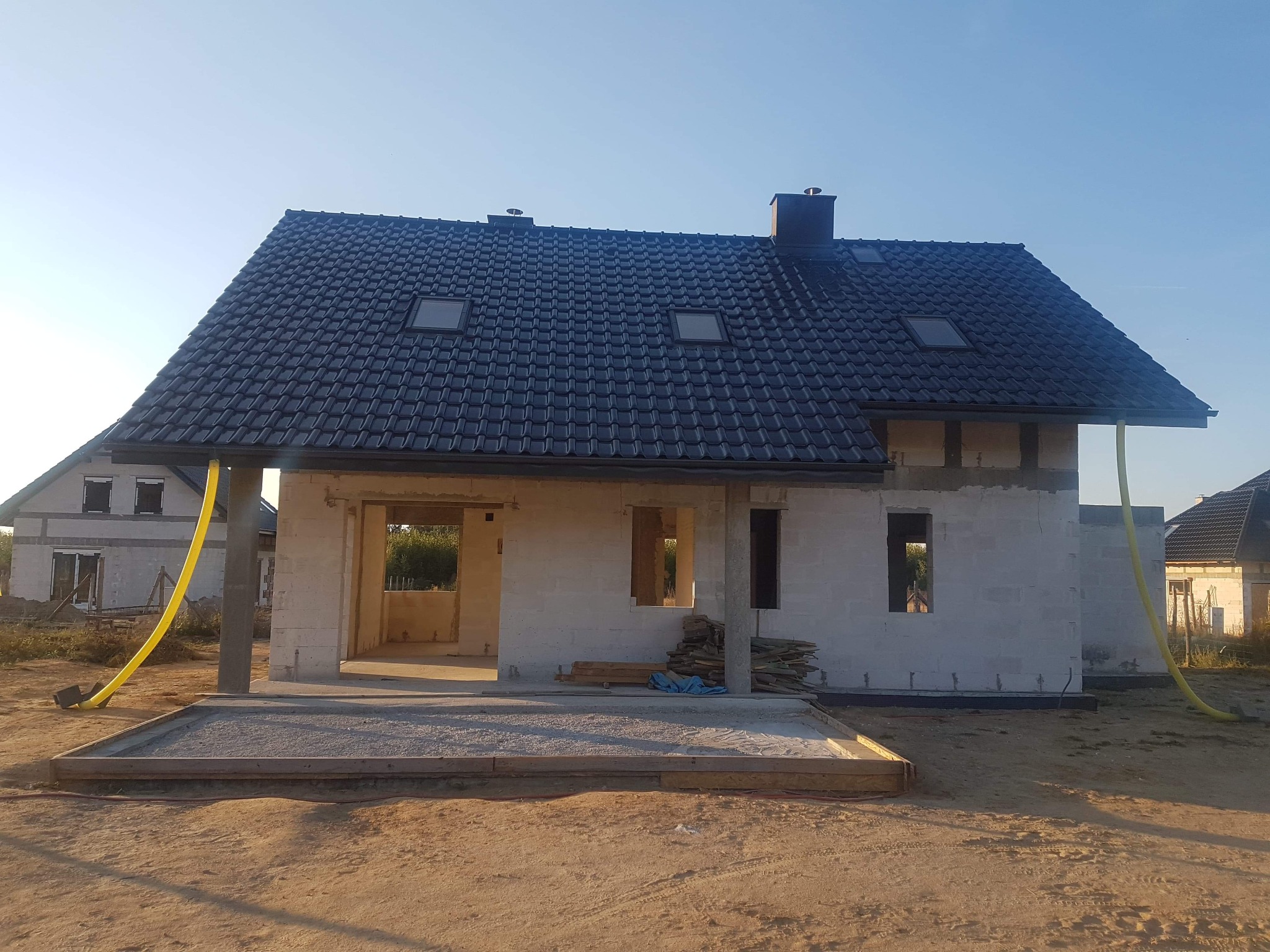 Dom w trakcie budowy z czarną dachówką, widoczne okna dachowe i komin, ściany z bloczków, żółte rury wentylacyjne na zewnątrz budynku.