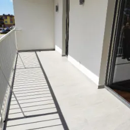 Jasny balkon z szarymi płytkami podłogowymi, białą balustradą i nowoczesnymi lampami ściennymi.