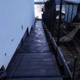 Ciemnoszara kostka brukowa ułożona wzdłuż białej ściany budynku i czarnego ogrodzenia panelowego, z widokiem na jezioro w tle.