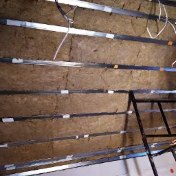 Skośny widok na poddasze w trakcie izolacji, widoczne metalowe profile, wełna mineralna, oraz fragment rusztowania i ściany z betonu komórkowego.