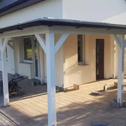 W trakcie budowy zadaszenia tarasu z białymi drewnianymi podporami i kostką brukową, widoczna ubijarka do gruntu.