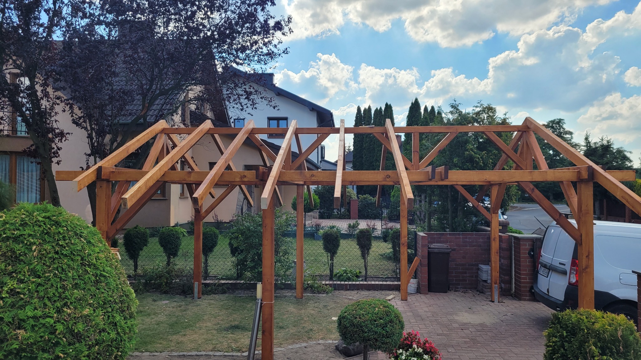 Drewniana konstrukcja pergoli w trakcie montażu, widoczna na tle zieleni ogrodu i zabudowań. Elementy konstrukcyjne z jasnego drewna, częściowo zmontowane.