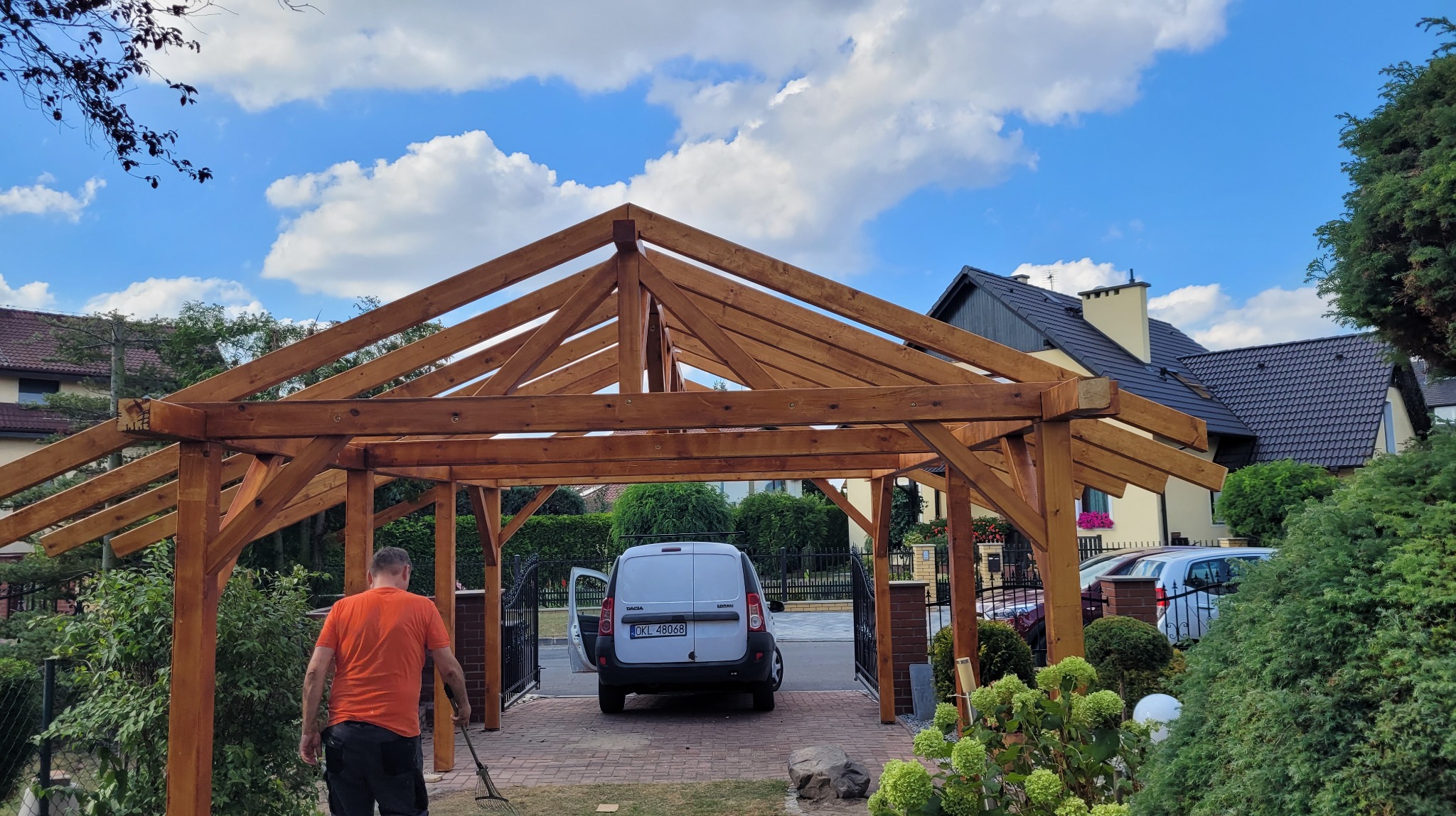 Drewniana konstrukcja z belek, prawdopodobnie wiata garażowa w trakcie budowy. Widoczny fragment posesji, samochód i osoba z grabiami.