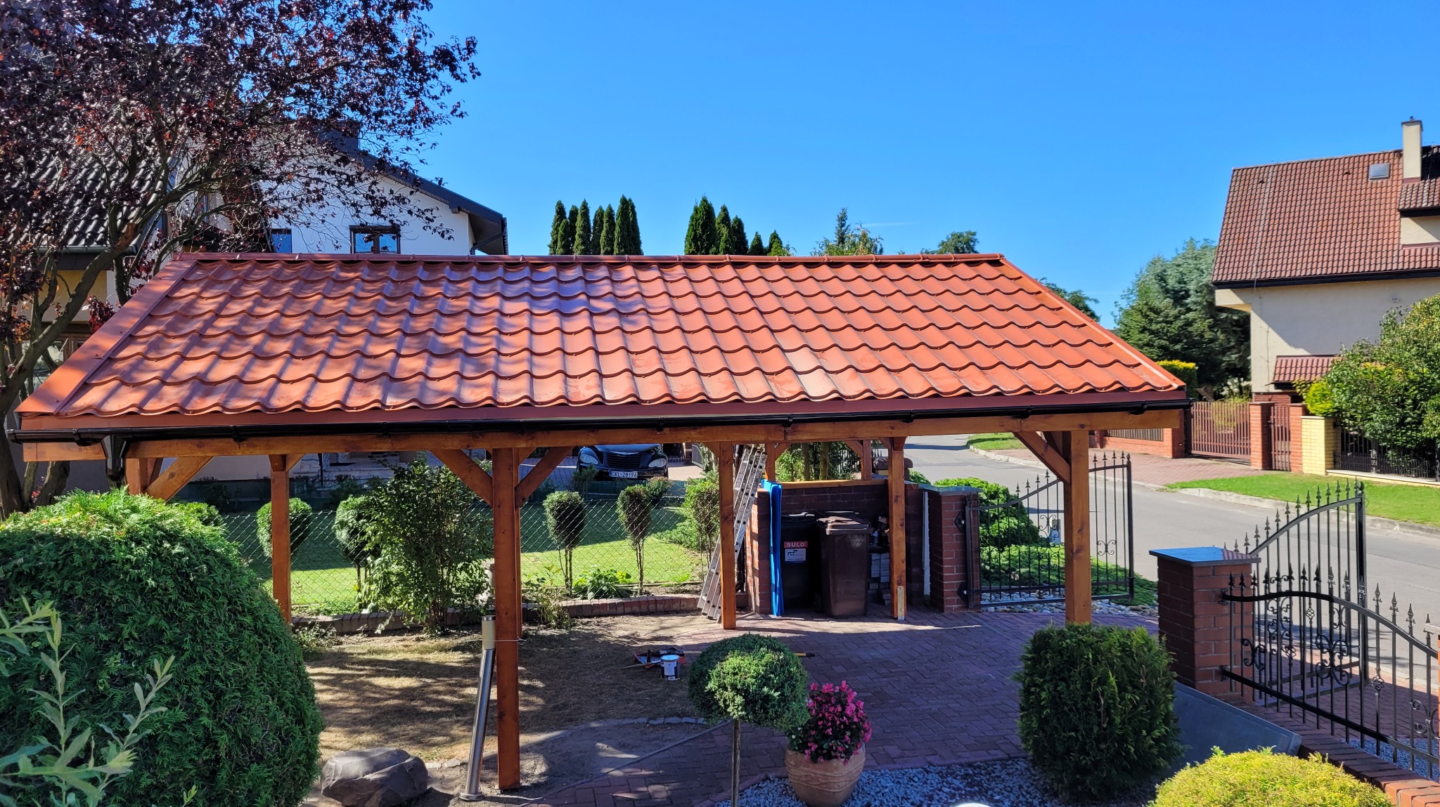 Drewniana wiata z czerwoną dachówką na posesji, widok z zewnątrz. W tle domy i zieleń, błękitne niebo. Konstrukcja z belek, widoczne detale wykończenia.