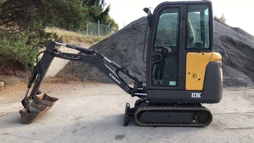 Żółto-szara mini koparka Volvo EC15C z łyżką, stojąca na betonowej nawierzchni, z hałdą gruzu w tle.