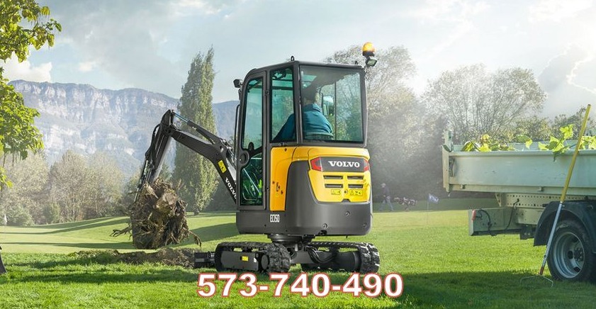 Żółto-czarna minikoparka Volvo EC15D z operatorem, podnosi korzeń drzewa z wykopu na trawniku; w tle góry i przyczepa z zielonymi gałęziami.