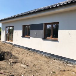 Kamil susel - Świeżo otynkowany dom jednorodzinny z brązowymi oknami, widoczna izolacja fundamentów i wykop w ziemi, sugerujący dalsze prace ziemne.