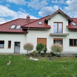 Kamil susel - Świeżo otynkowany dom jednorodzinny z czerwoną dachówką, widoczne okna dachowe, elewacja w kolorze złamanej bieli, dekoracyjny ogródek z młodymi drzewkami i rozłożony sprzęt budowlany na trawniku.