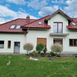 Świeżo otynkowany dom jednorodzinny z czerwoną dachówką, widoczne okna dachowe, elewacja w kolorze złamanej bieli, dekoracyjny ogródek z młodymi drzewkami i rozłożony sprzęt budowlany na trawniku.