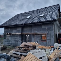 Kamil susel - Dom w trakcie ocieplania ścian zewnętrznych, widoczne rusztowania, stos materiałów budowlanych, ciemna elewacja, okna dachowe i pochmurne niebo.