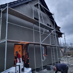 Kamil susel - Elewacja domu w trakcie szpachlowania, widoczne rusztowania, pracownik mieszający zaprawę, radio budowlane Makita na workach zaprawy, pochmurne niebo.