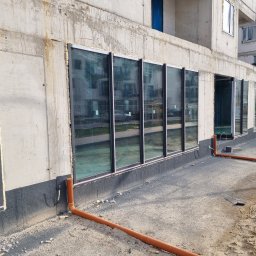 Kamil susel - Montaż okien w surowym budynku z widoczną izolacją fundamentów i rurami odprowadzającymi wodę, pianka montażowa wokół ram okiennych.