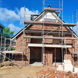 Kamil susel - Budynek w trakcie murowania z cegły, otoczony rusztowaniami, z widocznym stosem cegieł na pierwszym planie i błękitnym niebem w tle.