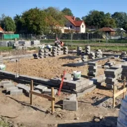 Wykop pod fundamenty z ułożonym szalunkiem i częściowo wymurowanymi bloczkami betonowymi, widoczne zapasy bloczków i pracownicy w oddali.